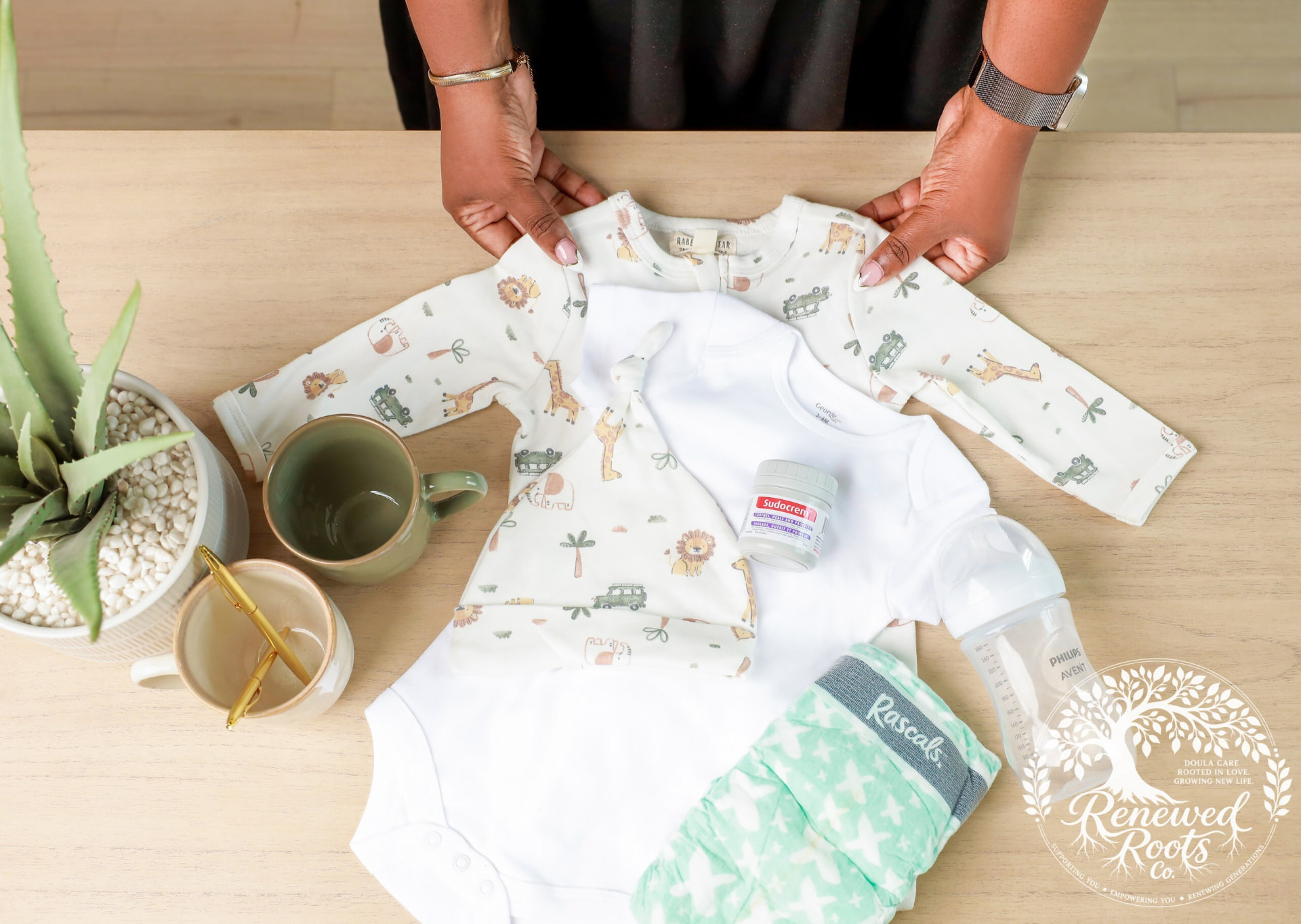 Hands holding a safari-print baby sleeper above newborn essentials, including a bottle and care items